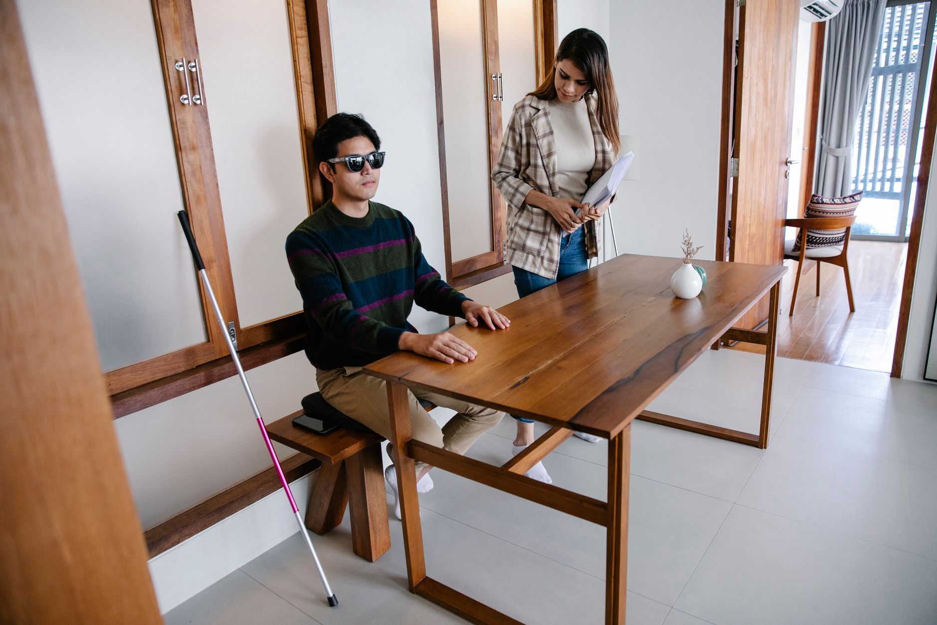 A visually impaired man sits at a wooden table with a supportive woman nearby in a modern home.