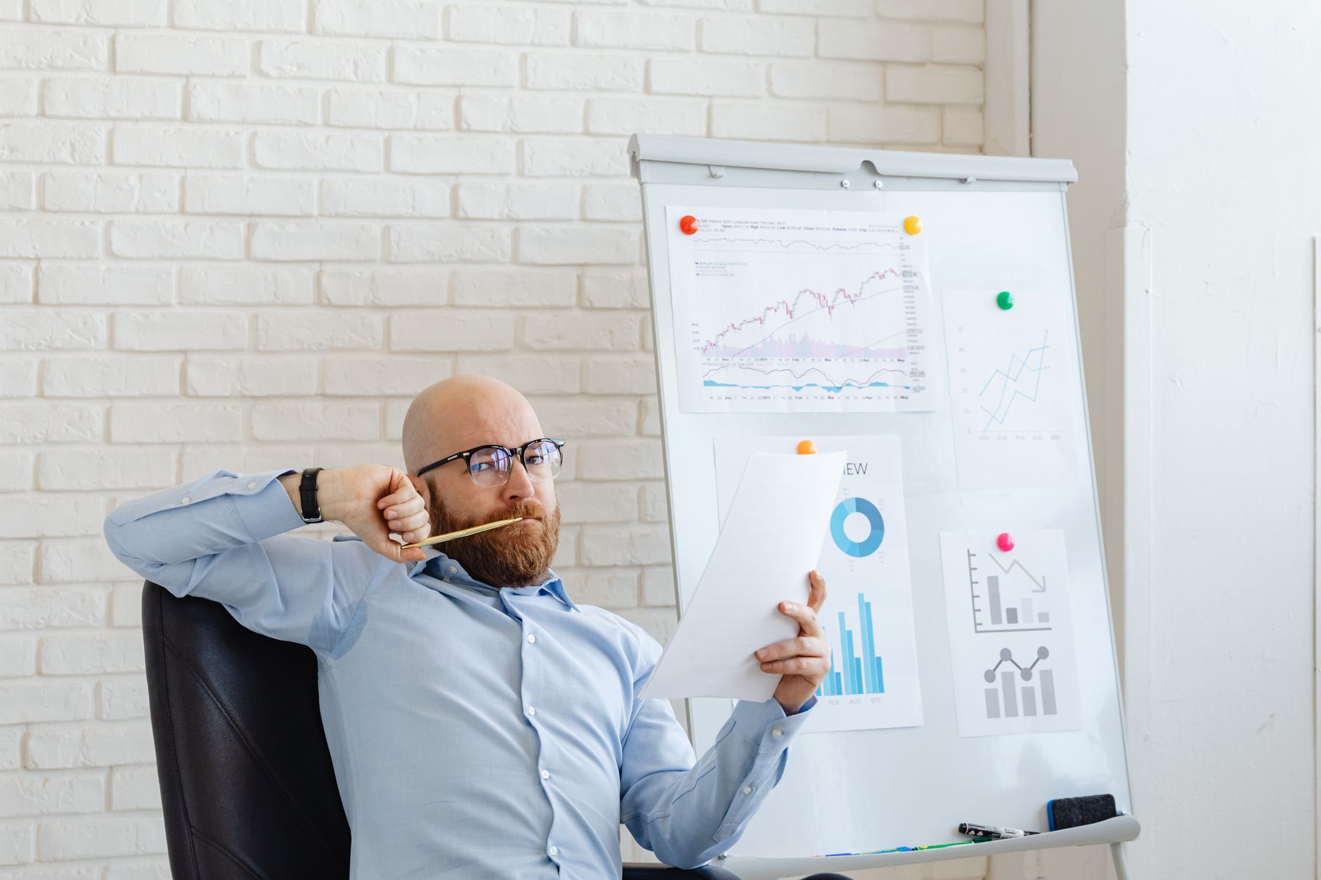 A businessman in glasses reviews financial charts, deep in thought by a whiteboard with graphs.