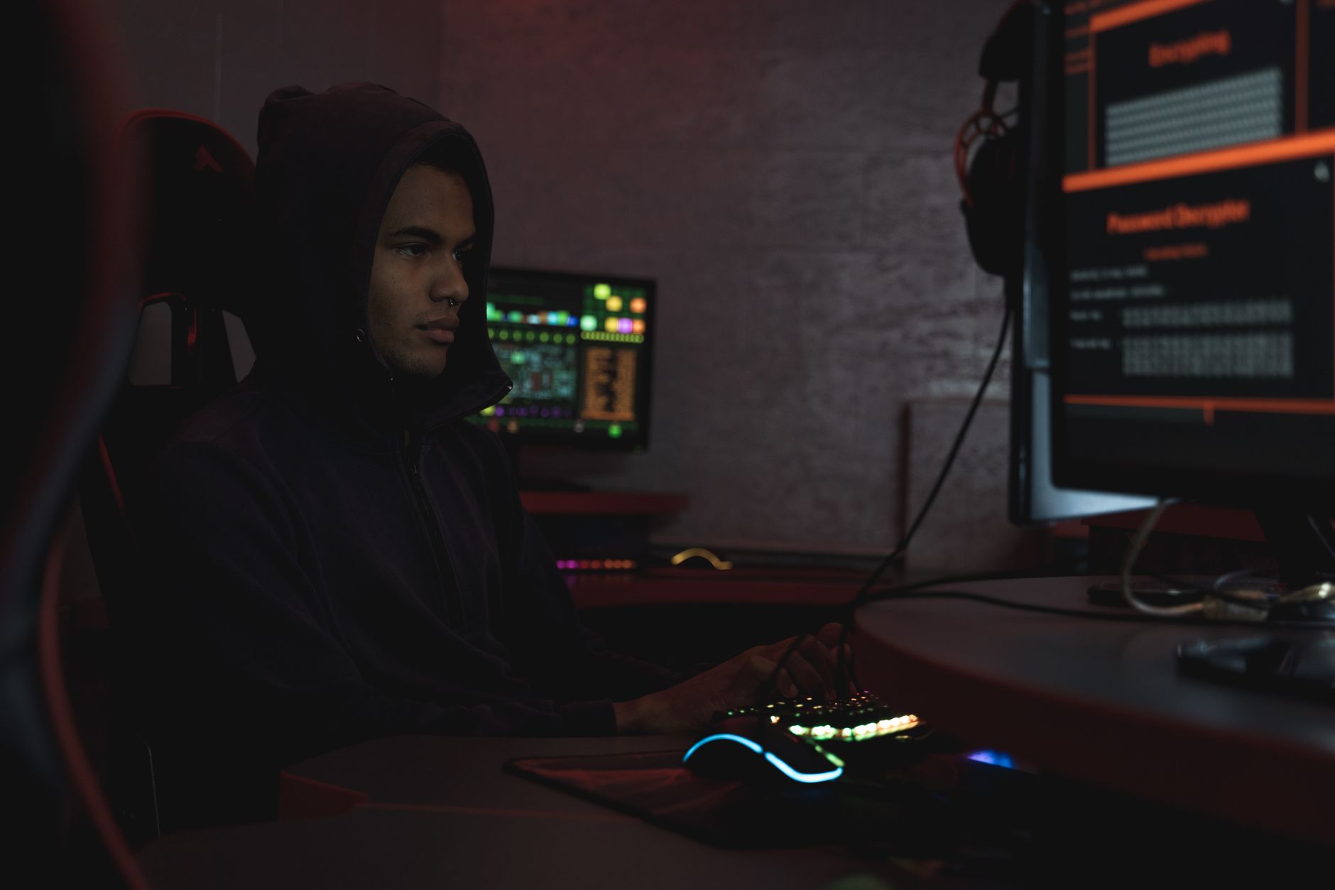 A hacker in a hoodie working in a dimly lit room, focusing on cyber security tasks on multiple monitors.