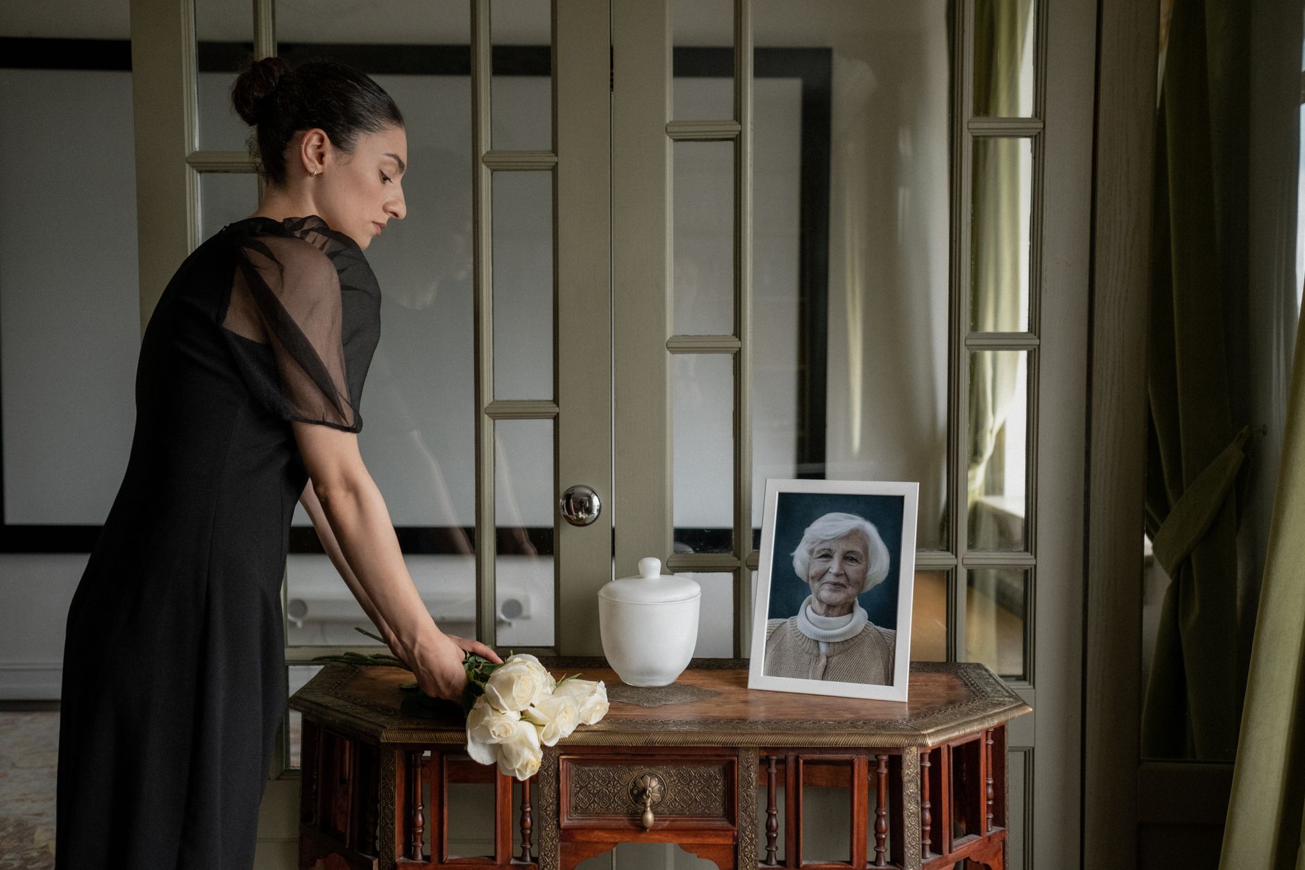 A woman in black holds flowers near an urn and framed photo, conveying mourning.