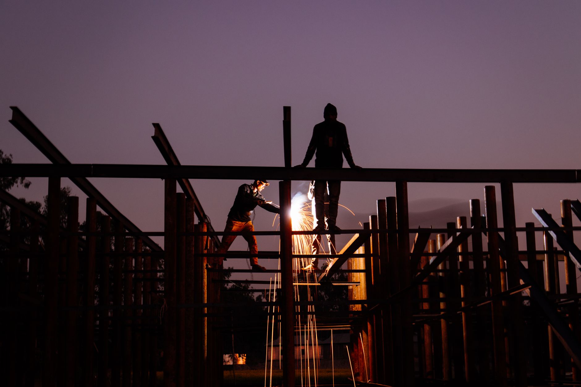 Silhouettes of welders working at a construction site during sunset, creating dramatic sparks.